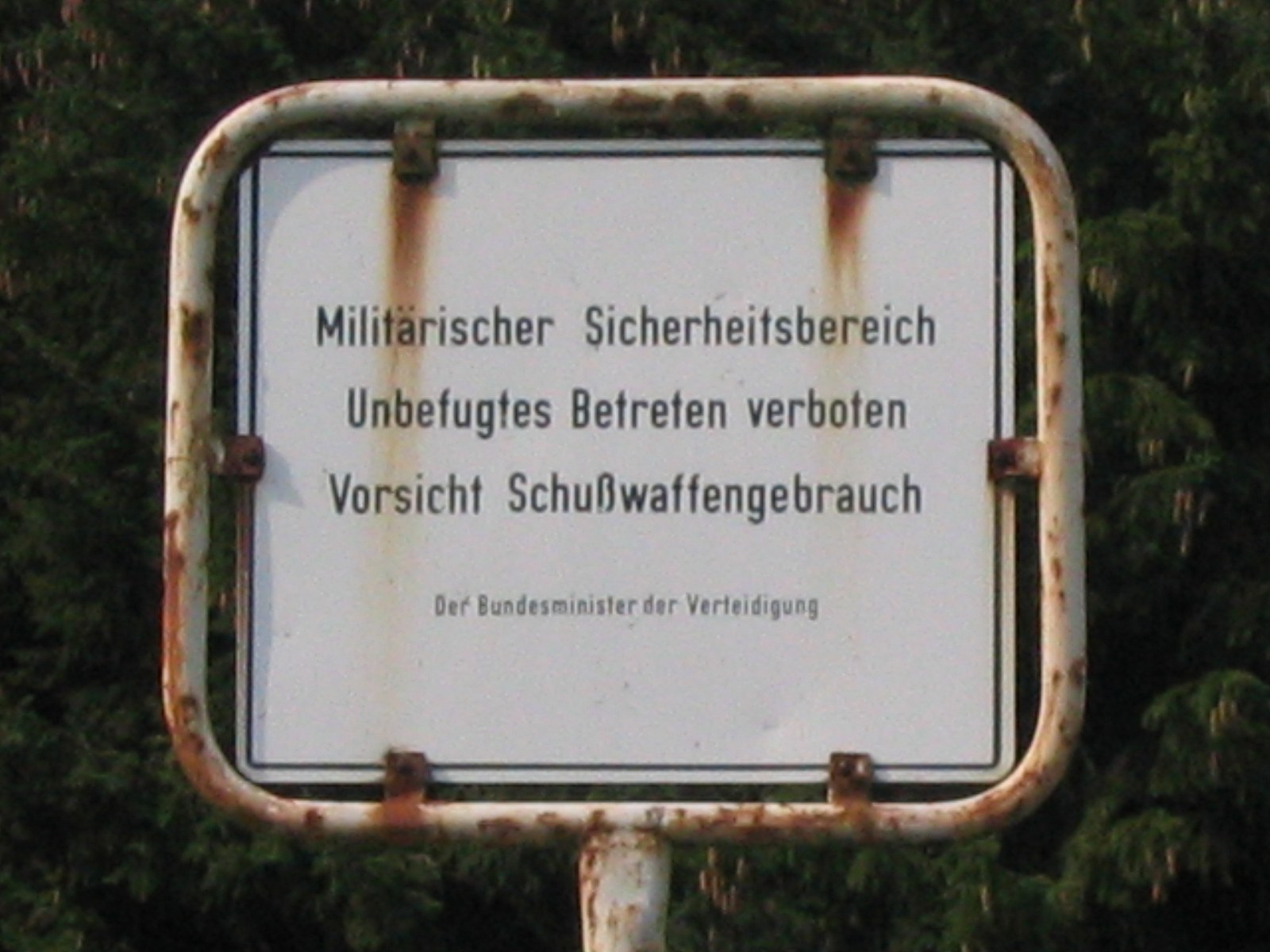 Ein angerostetes Schild vor Nadelbäumen: Militärischer Sicherheitsbereich, Unbefugtes Betreten verboten, Vorsicht Schußwaffengebrauch