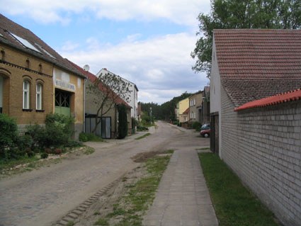 Eine sandige Dorfstraße mit Gehweg, links ein Haus aus gelben Klinkern, rechts eine helle Mauer unter roten Dachziegeln
