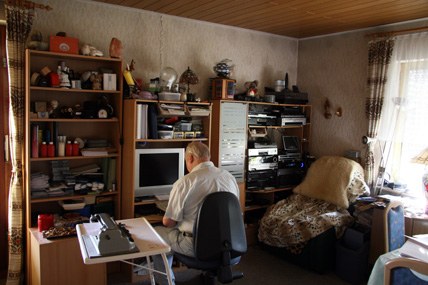 Ein Wohnraum voller Regale, Hi-Fi-Geräte und Bildschirm, Josef Arens sitzt mit dem Rücken zur Kamera